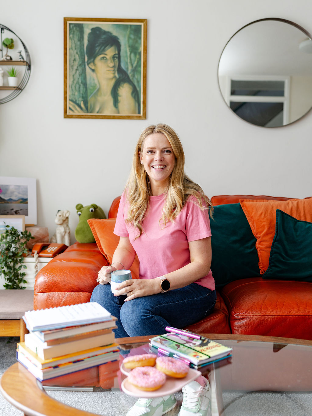Author and entrepreneur Becky Wash sits on her sofa smiling with a cup of coffee . A plate of doughnuts sit amongst her pile of books on the coffee table in front of her.